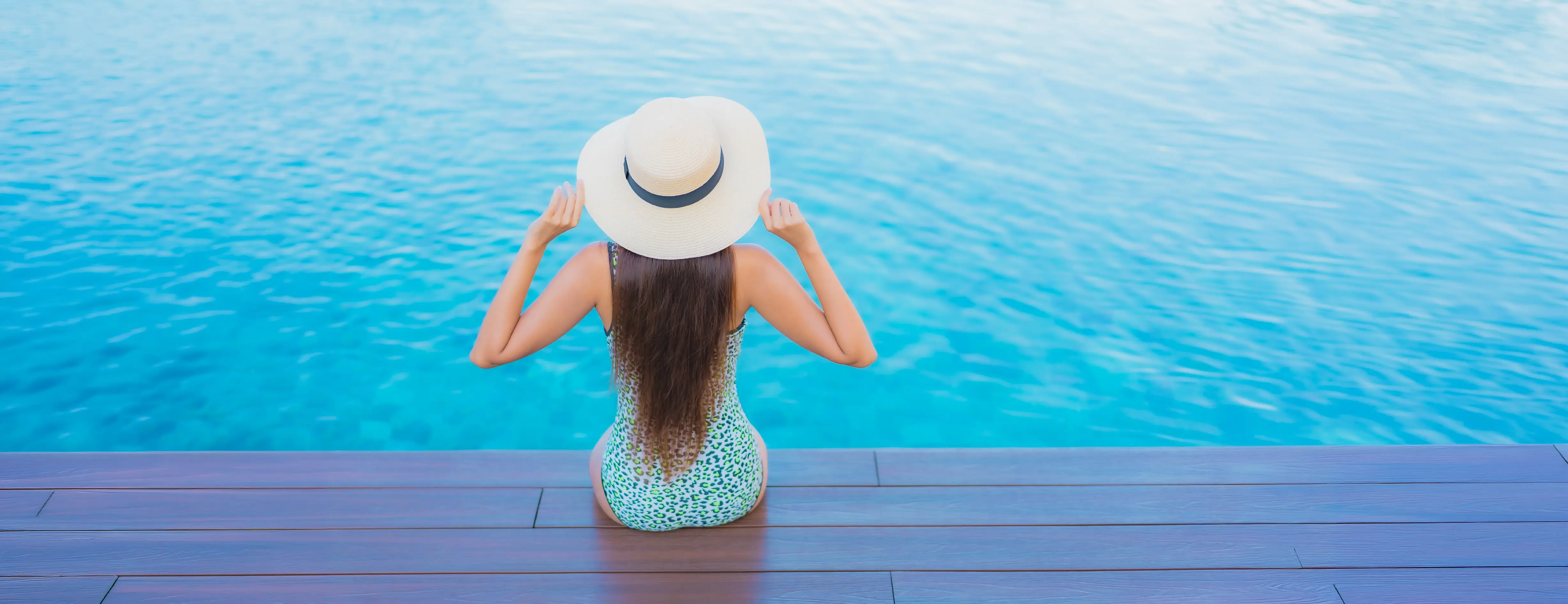 A woman in a sun hat sits at the edge of a still blue pool.